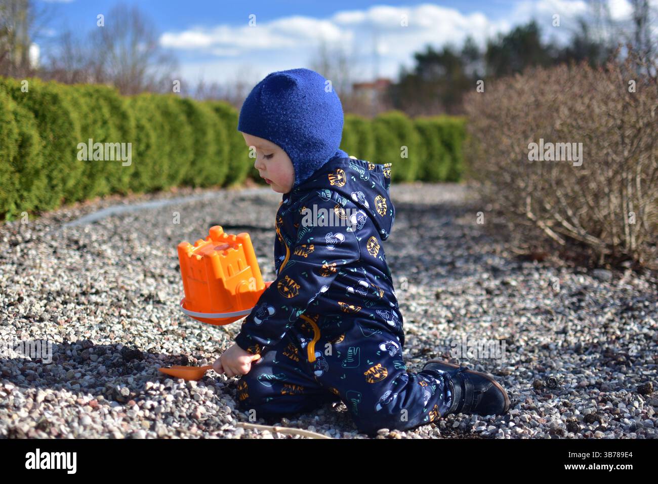 Garçon tout-petit joue avec un seau et une pelle dans le parc public. Enfant creusant des cailloux avec des jouets de sable à l'extérieur au printemps. Banque D'Images