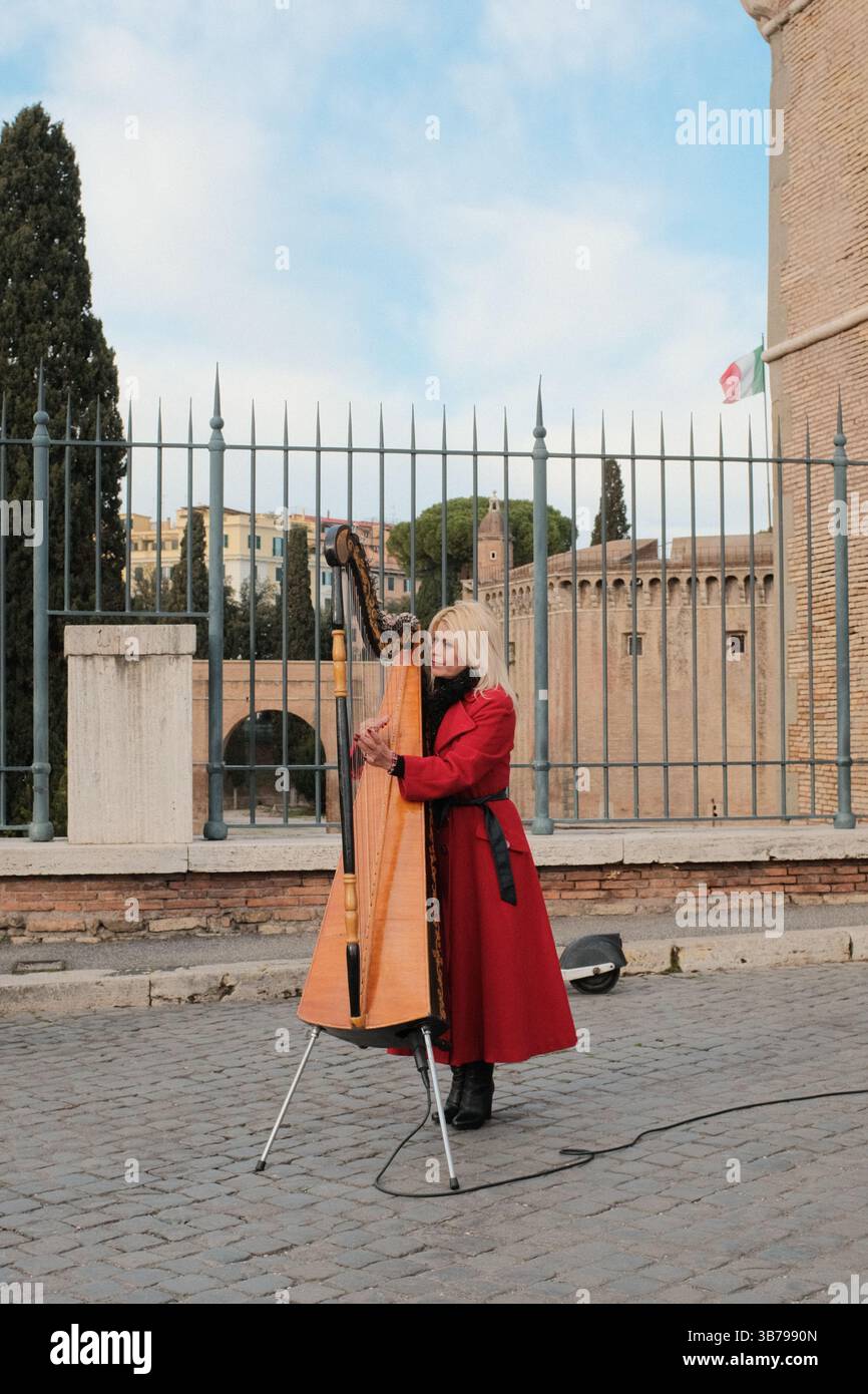 Femme en manteau rouge jouant de la harpe dans une rue pavée près de Castel Sant Angelo à Rome jouant de la musique classique à l'extérieur sous le ciel du jour. Photo de haute qualité Banque D'Images