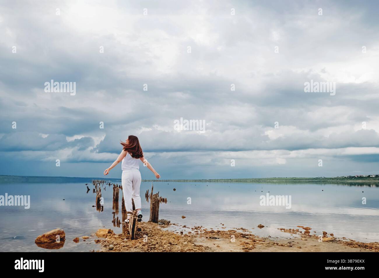 Femme marche sur des poteaux en bois par l'eau calme sereine sous un paysage de nuages dramatique Banque D'Images