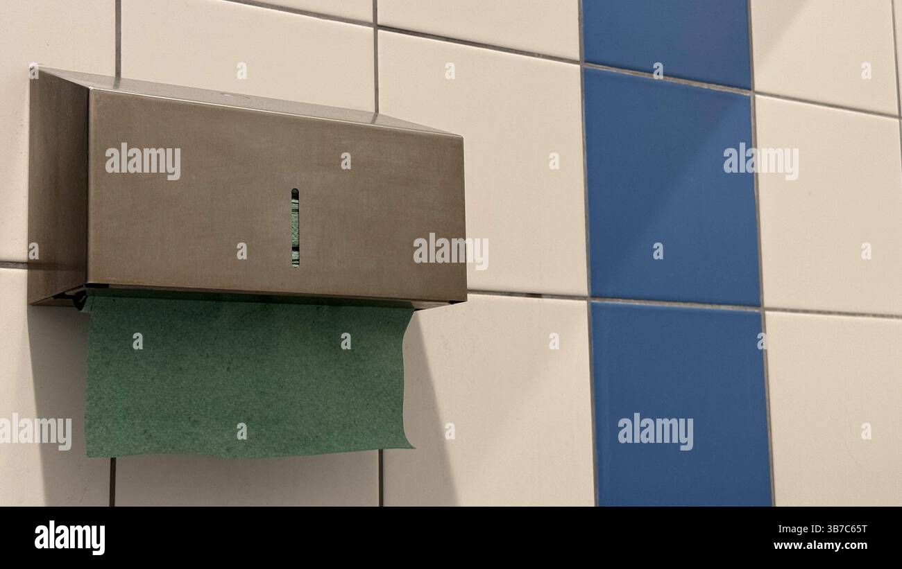 Distributeur d'essuie-mains en papier en acier inoxydable avec serviette verte contre le mur des toilettes carrelé blanc et bleu capture un design minimaliste et une utilité. Banque D'Images
