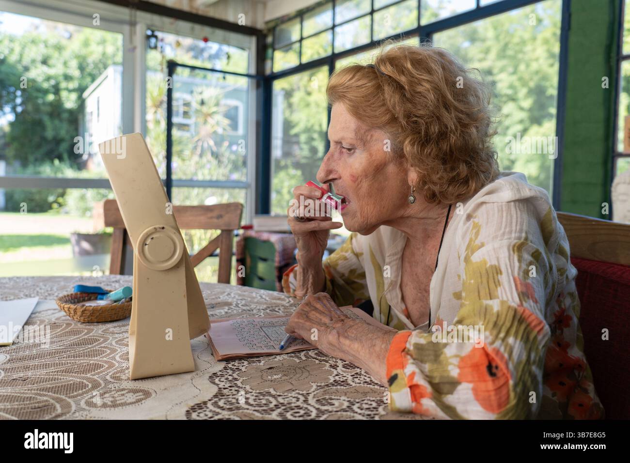 Femme âgée assise à une table dans sa maison confortable, appliquant du rouge à lèvres rouge avec un petit miroir, embrassant sa routine de beauté et se préparant en toute confiance fo Banque D'Images