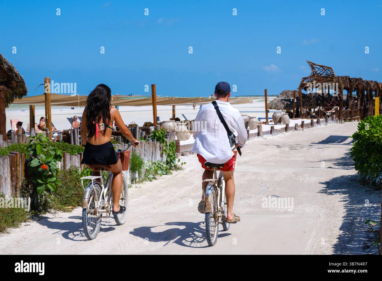 Île Holbox, Mexique – 10 février 2025 : un couple fait du vélo sur un sentier côtier sablonneux près des bars de plage et des chaises longues sur Isla Holbox, une île sans voiture Banque D'Images