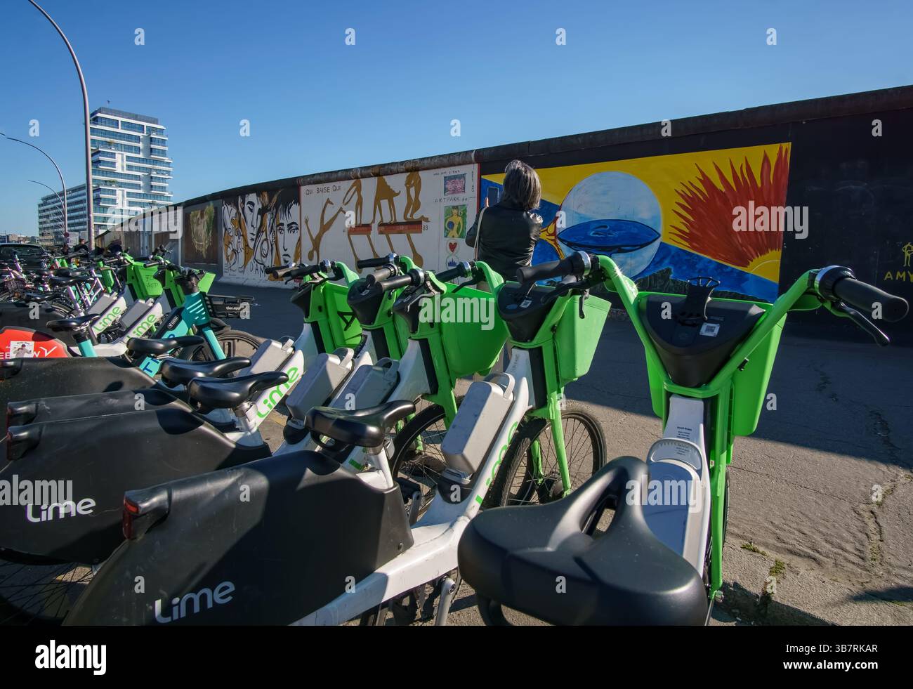 Des vélos lime sont alignés à East Side Gallery tandis qu'une femme prend des photos de peintures murales sur le mur historique de Berlin sous le soleil du printemps. Banque D'Images