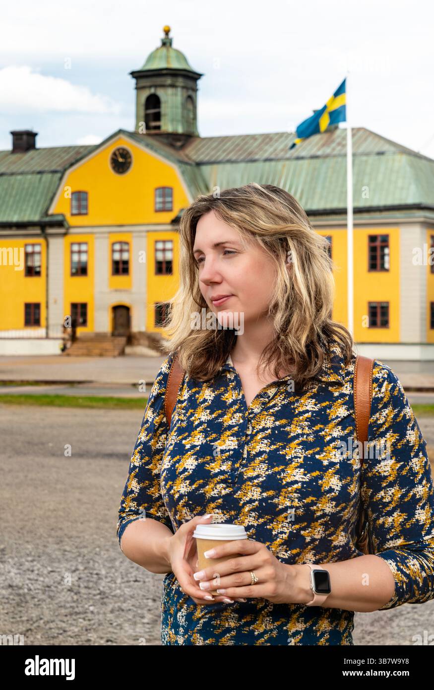 Femme se tient avec une tasse à café, ornée de clous élégants, bague de mariage et montre moderne, en face d'un bâtiment jaune suédois historique Banque D'Images
