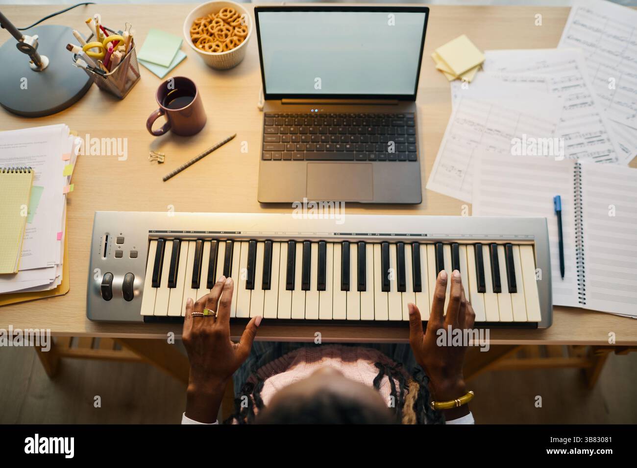Vue aérienne d'une femme composant de la musique en jouant au clavier au bureau avec des feuilles de musique, tasse à café et ordinateur portable. Espace de travail soigné aménagé pour la création musicale Banque D'Images