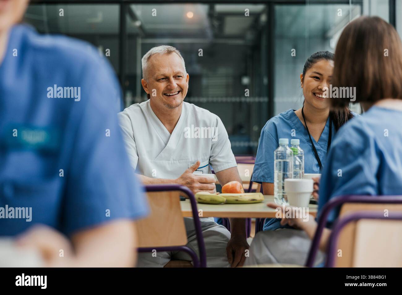 Médecin masculin souriant parlant avec des collègues tout en prenant le petit déjeuner à table dans la cafétéria de l'hôpital Banque D'Images