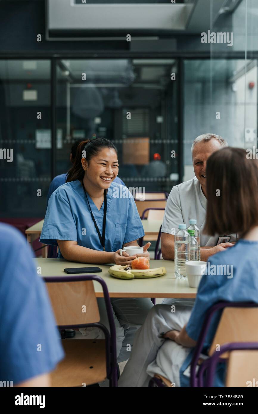Heureuse jeune femme professionnelle médicale prenant un café tout en parlant et assis avec des collègues à table à l'hôpital CAF Banque D'Images
