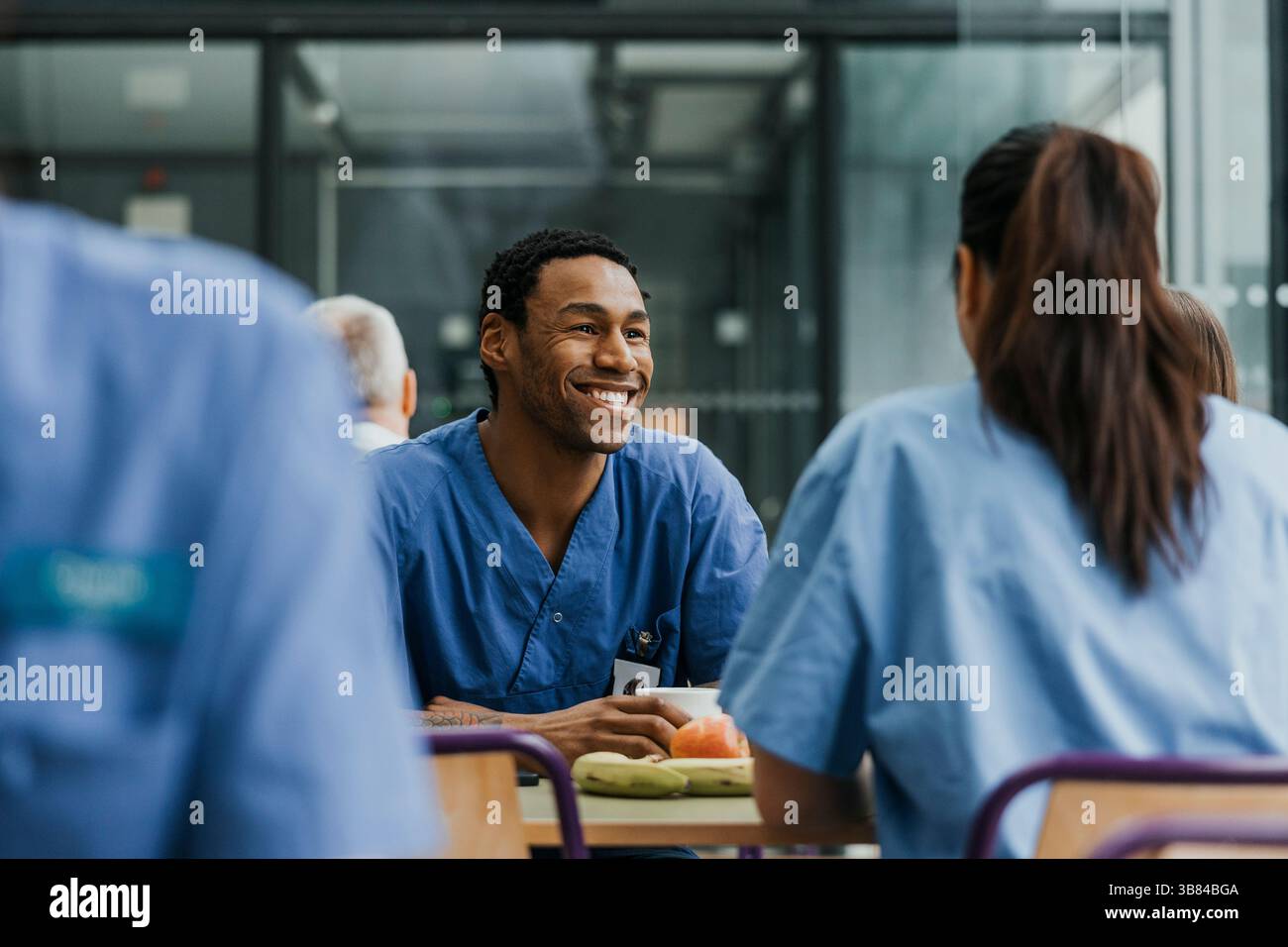 Heureux professionnel médical masculin prenant le café tout en parlant et assis avec des collègues à table dans la cafétéria de l'hôpital Banque D'Images