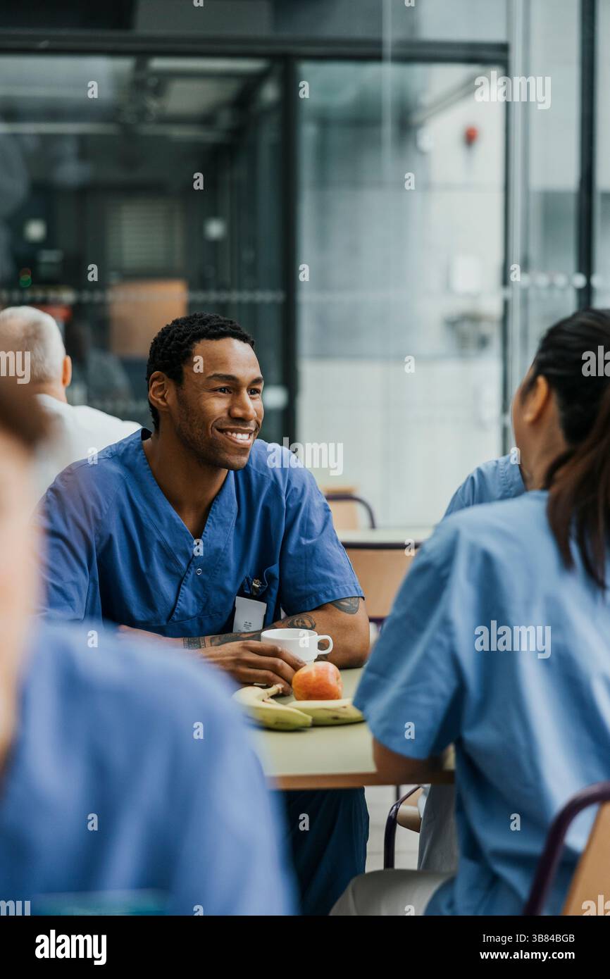 Professionnel médical masculin souriant prenant un café tout en étant assis avec des collègues à table dans la cafétéria de l'hôpital Banque D'Images