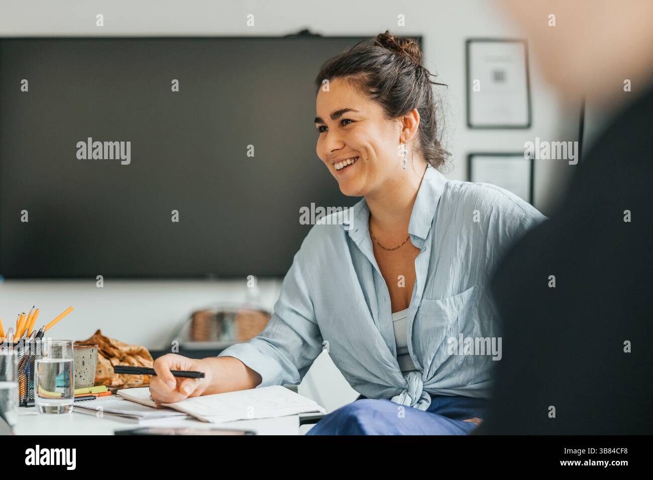 Heureuse jeune femme d'affaires écrivant dans le journal tout en parlant avec un collègue au bureau Banque D'Images