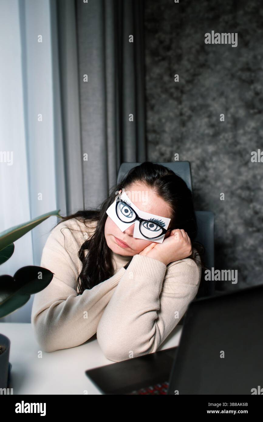 Concept de surmenage au bureau. Pas le temps de dormir. Travailleuse de bureau avec des yeux peints sur des autocollants imitant le travail devant l'ordinateur pendant le sommeil Banque D'Images