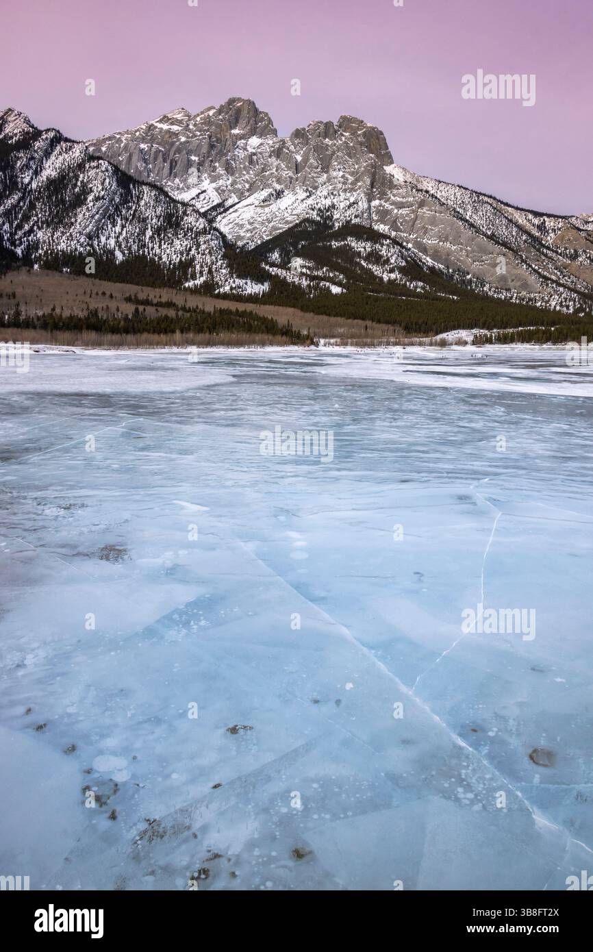 Un lac gelé calme s'étend au premier plan, présentant des configurations de glace complexes sous un ciel crépusculaire. Des montagnes majestueuses s'élèvent en arrière-plan, Banque D'Images