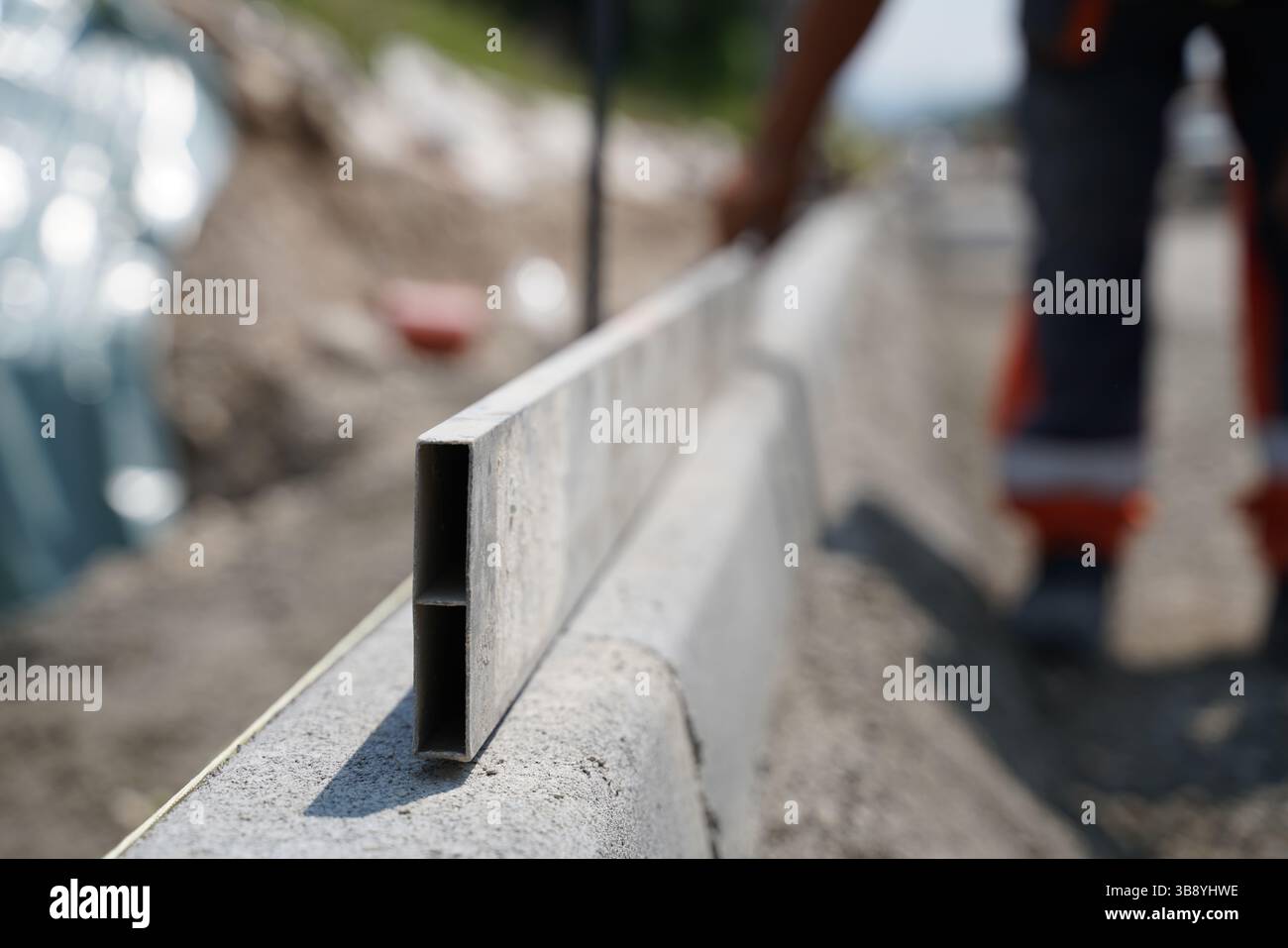 Gros plan d'un travailleur utilisant une barre de mise à niveau en aluminium pour vérifier le niveau de bordure de la route sur le chantier de construction d'autoroute (travailleur flou en arrière-plan) Banque D'Images