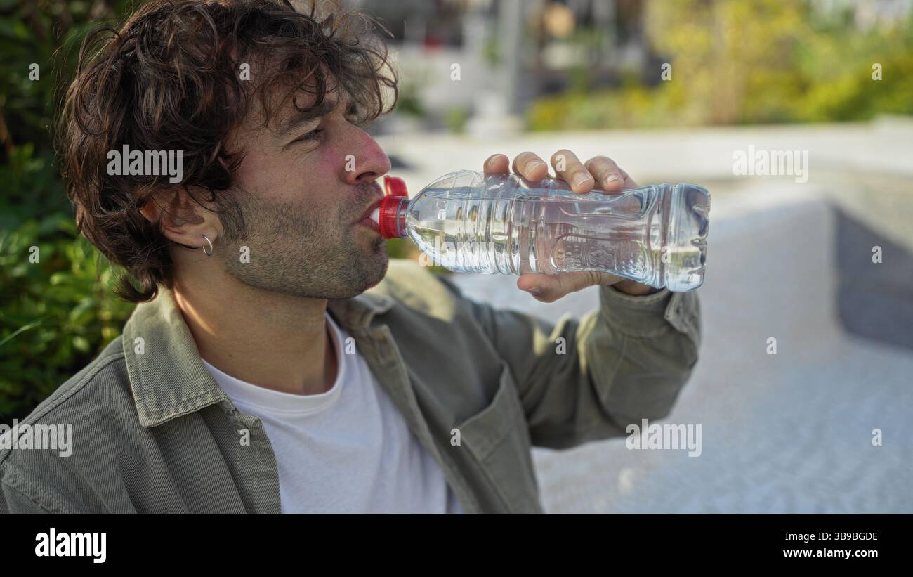 Jeune homme buvant de l'eau d'une bouteille dans un parc urbain, mettant en valeur un style décontracté et rafraîchissant scène en plein air avec de la verdure et du soleil dans le dos Banque D'Images