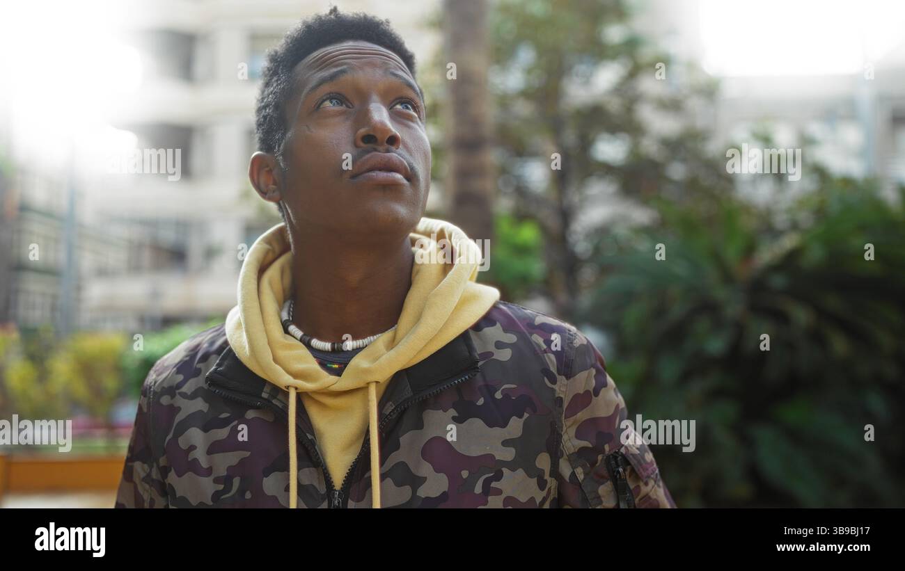 Jeune homme dans le parc urbain regarde avec une expression réfléchie, entouré de verdure et de paysage urbain, portant un sweat à capuche décontracté et veste camouflage suggérant un Banque D'Images