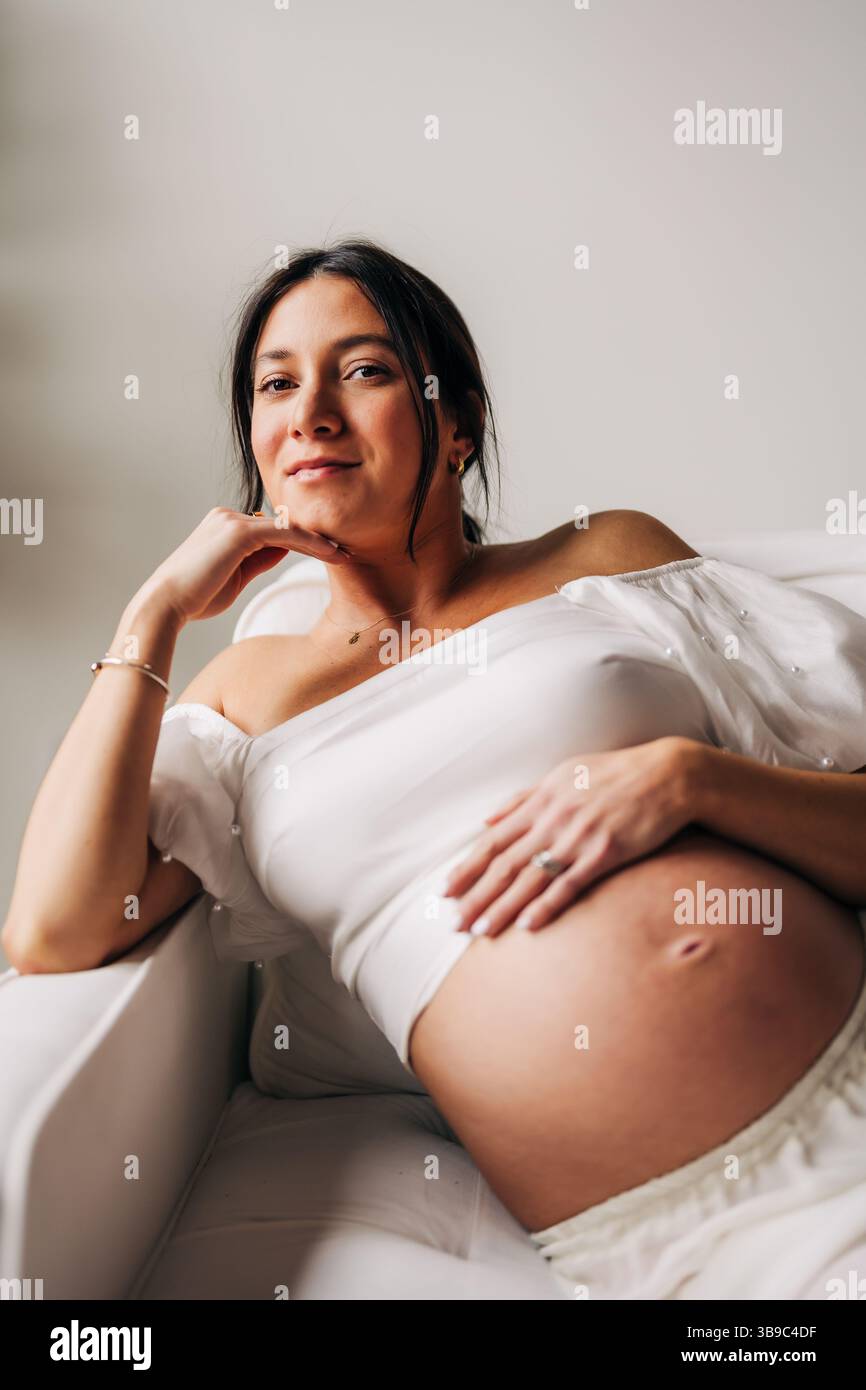 Femme enceinte se prélassant dans une tenue blanche avec la main sur le ventre, souriant Banque D'Images