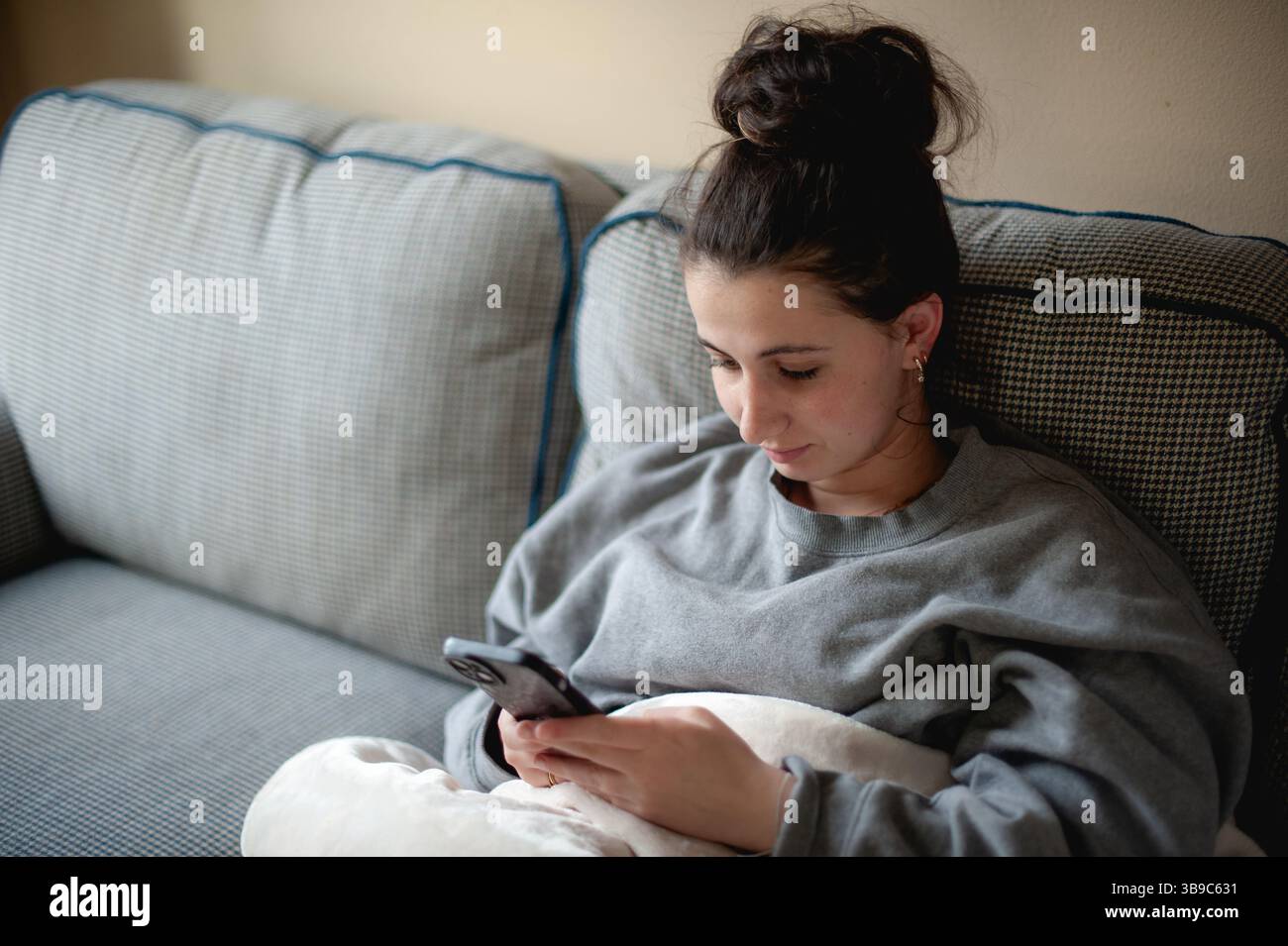 Adolescent se relaxant sur le canapé à l'aide d'un smartphone tout en enveloppant dans une couverture douce. Banque D'Images