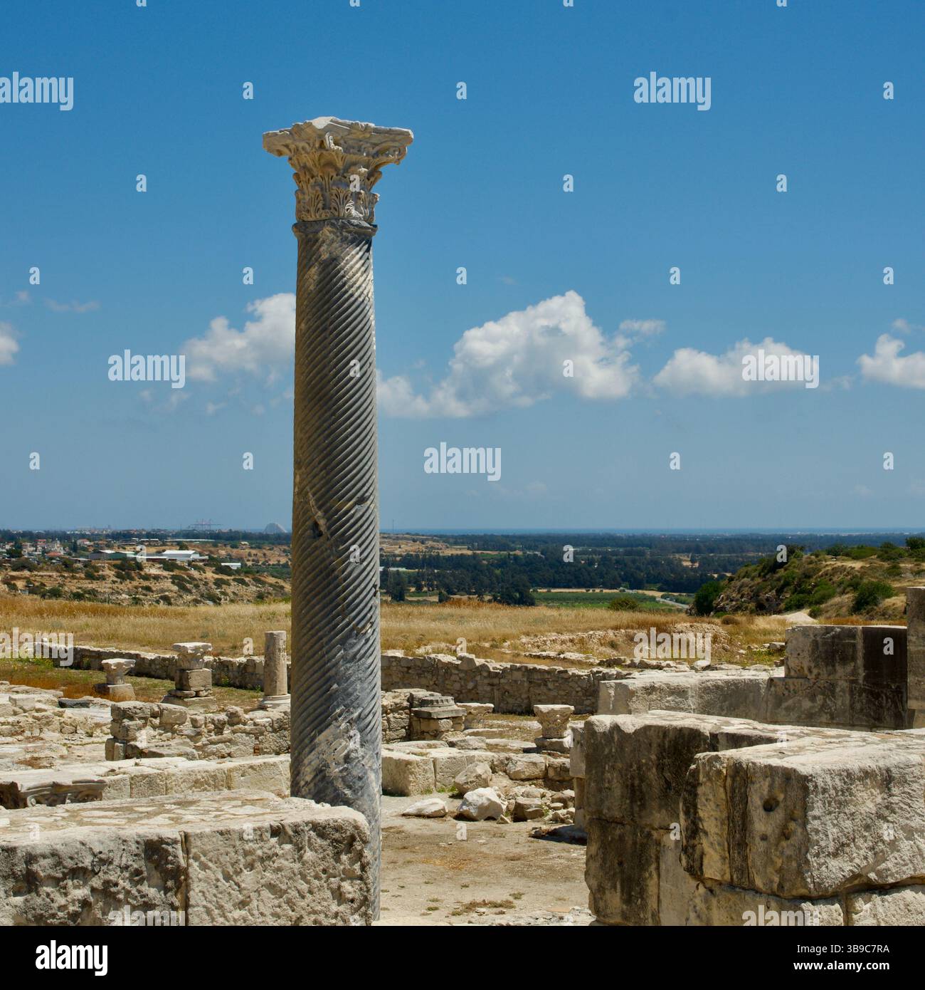 Ancienne colonne de style corinthien entourée de ruines ensoleillées dans un paysage rural serein, symbolisant l'histoire. Banque D'Images