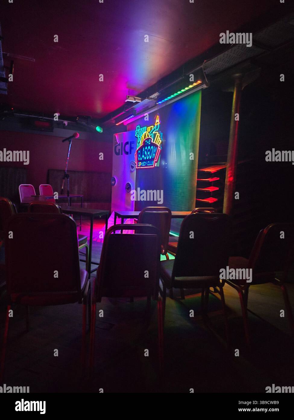 Une scène de club de comédie faiblement éclairée avec un pied de microphone, des chaises disposées pour le public et un panneau lumineux qui indique « GICF » et « Comedy Club ». Banque D'Images