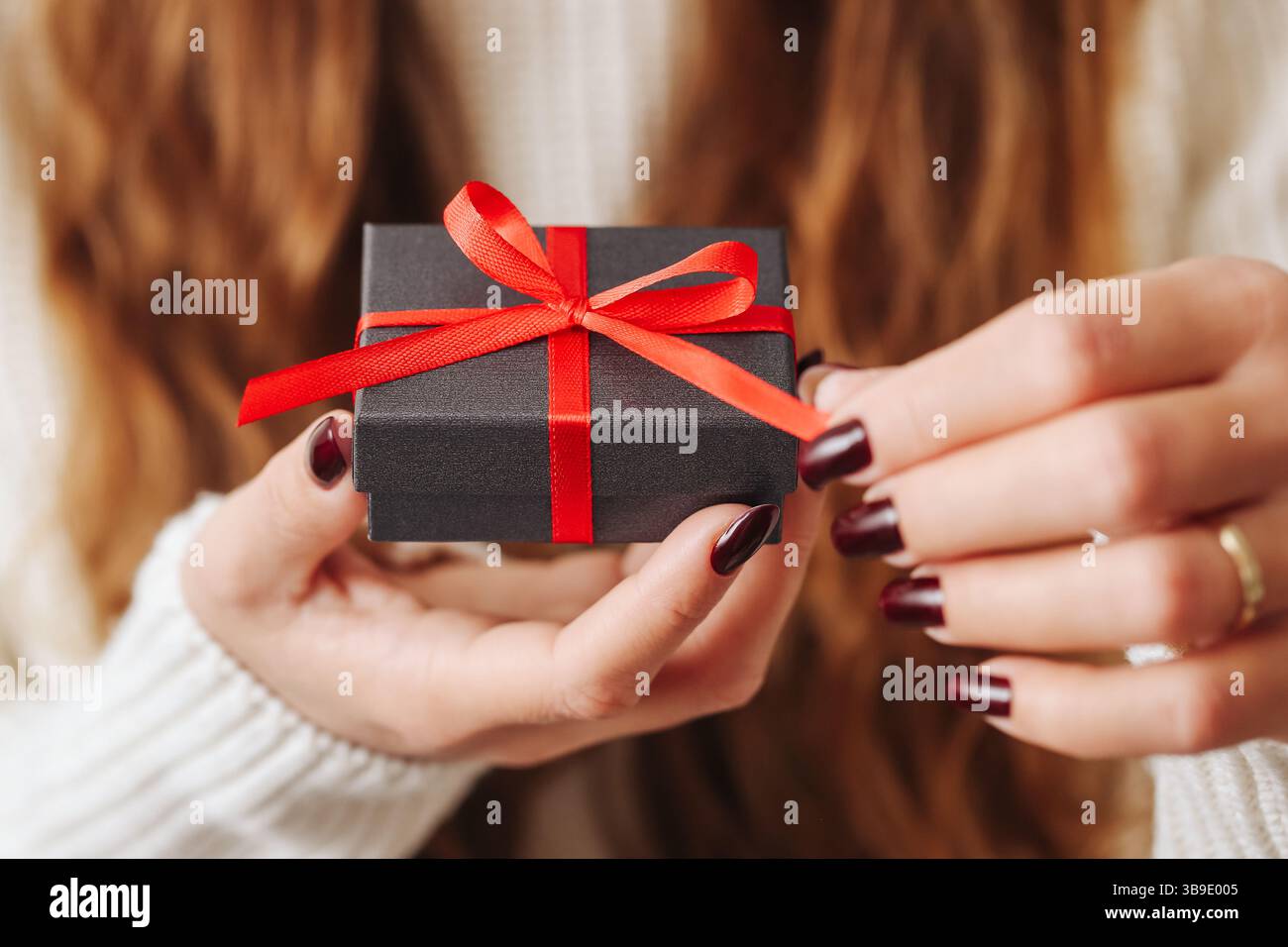 Boîte cadeau élégante moment. Petit paquet noir avec ruban rouge. Les mains de la femme délirent l'arc. Ambiance festive. Concept de don présent. Banque D'Images