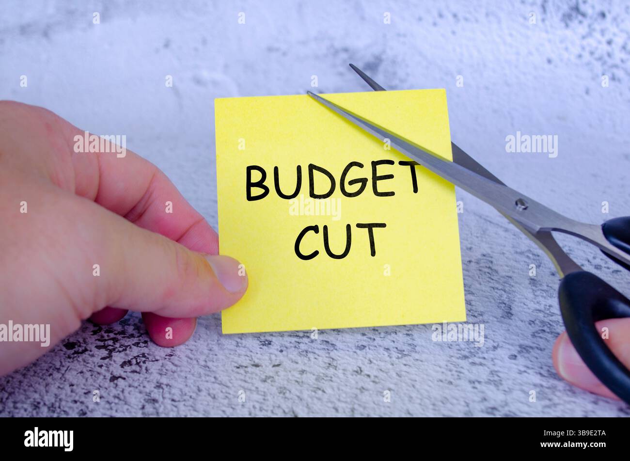 Main tenant une note collante jaune avec les mots budget Cut écrit dessus, tandis qu'une paire de ciseaux coupe à travers la note Banque D'Images