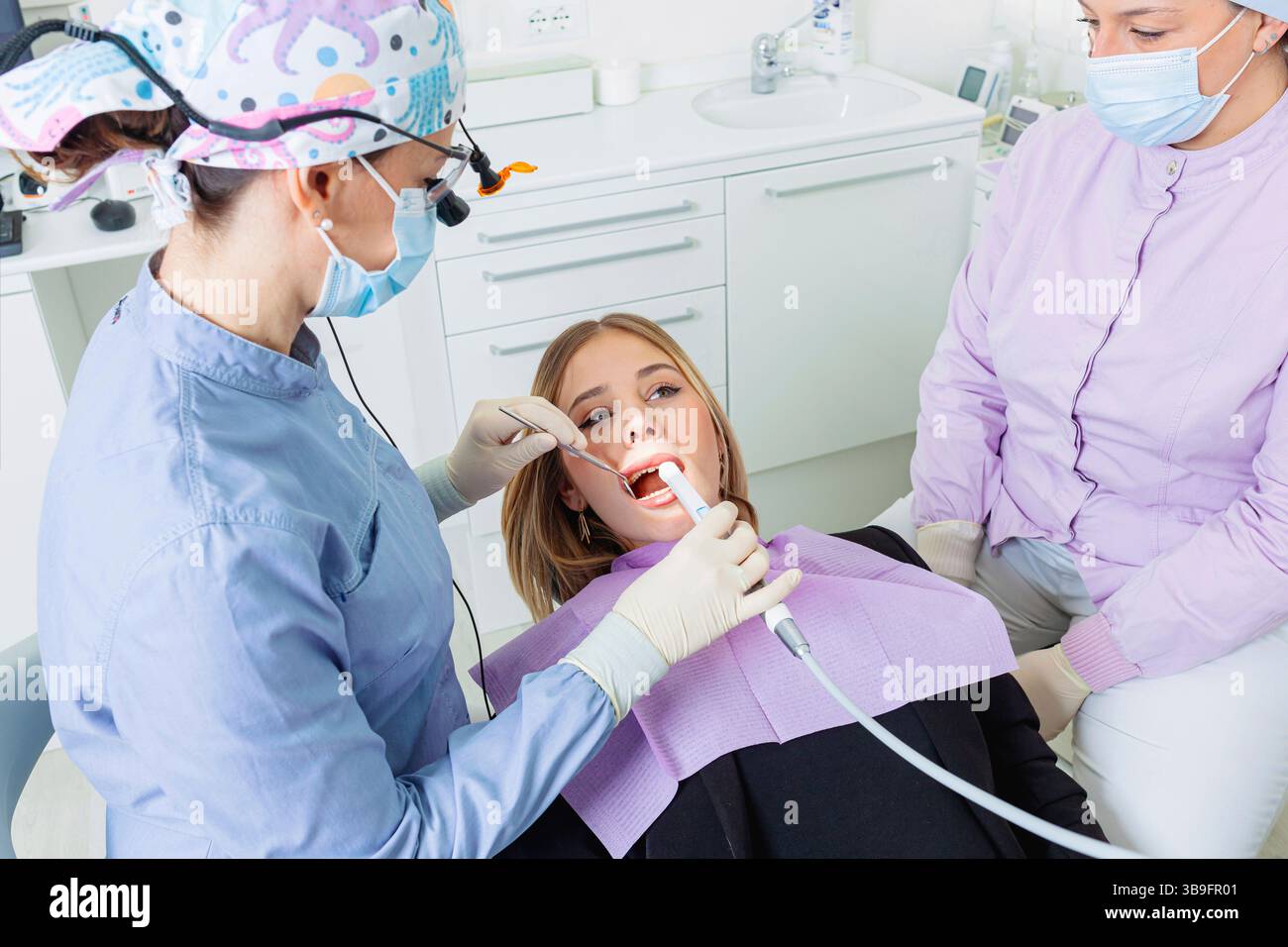 Dentiste portant des loupes chirurgicales effectuant un examen dentaire sur une jeune femme avec équipement dentaire dans une clinique dentaire Banque D'Images