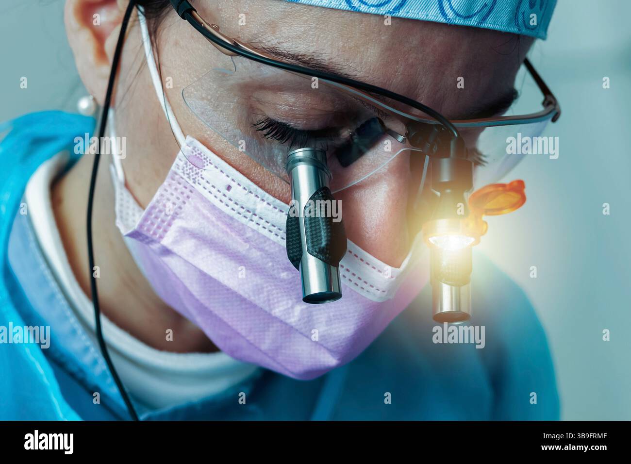 Dentiste femme portant une casquette chirurgicale, des loupes avec lumière et un masque rose, effectuant un examen dentaire approfondi dans un cadre de clinique dentaire moderne Banque D'Images