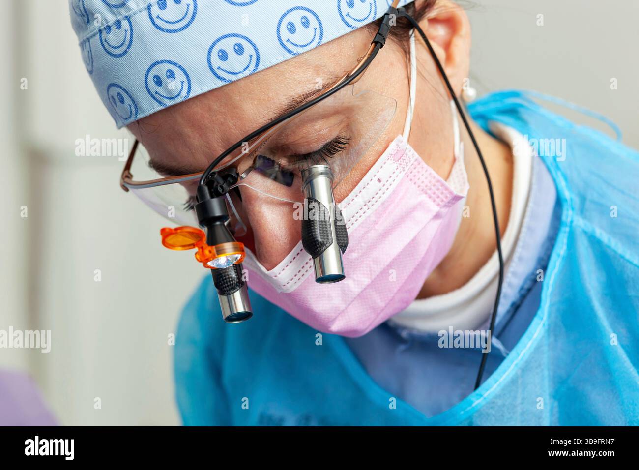 Dentiste femme portant une casquette chirurgicale, des loupes avec lumière et un masque rose, effectuant un examen dentaire approfondi dans un cadre de clinique dentaire moderne Banque D'Images