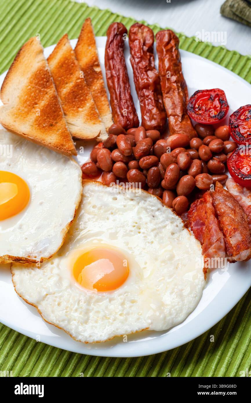 Il sert deux œufs frits accompagnés de saucisses, de bacon croustillant, de haricots et de tranches de pain grillé, créant ainsi un copieux petit déjeuner anglais complet sur une assiette blanche immaculée Banque D'Images