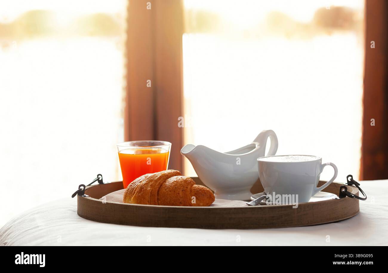 Plateau en bois avec cappuccino, croissant et jus d'orange reposant sur des draps blancs dans une chambre d'hôtel ensoleillée, offrant une expérience de petit déjeuner confortable et invitante Banque D'Images