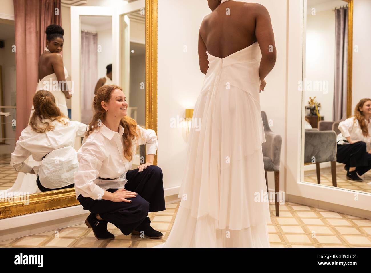 Une mariée à être porte une robe blanche superposée, debout dans une cabine d'essayage. Sa créatrice, dans une chemise blanche, l'assiste, assurant un ajustement parfait. La chambre ha Banque D'Images