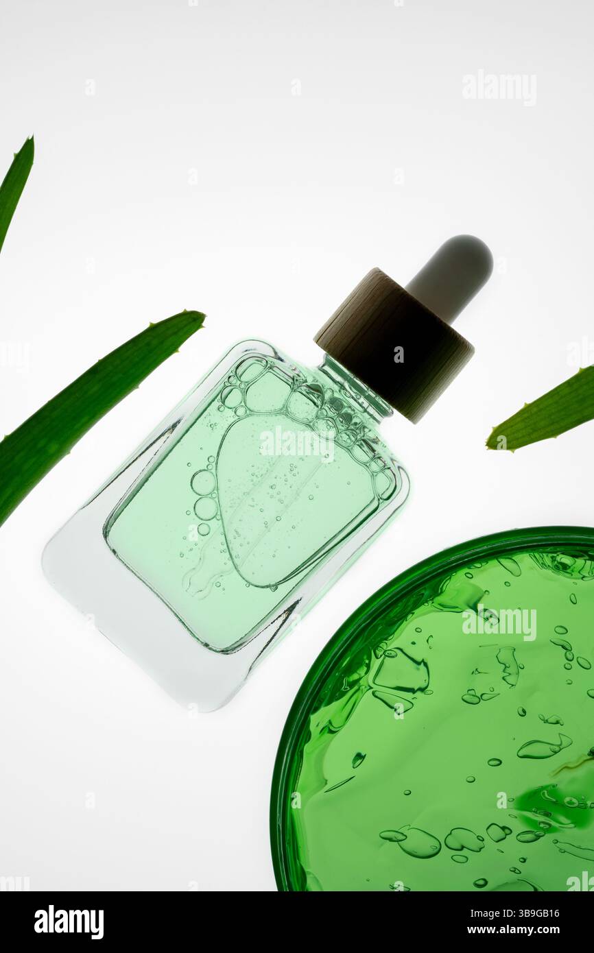 Une bouteille de sérum d'aloe vera avec un compte-gouttes repose sur un fond blanc, entouré de feuilles vertes. L'arrangement minimaliste évoque un sens de natura Banque D'Images