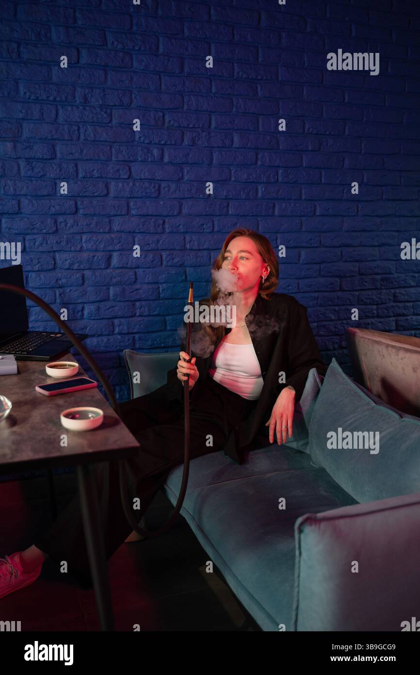 Une femme aime une séance de fumer détendue, assise sur un canapé bleu avec un narguilé. L'éclairage ambiant rouge et bleu crée une atmosphère apaisante. Banque D'Images