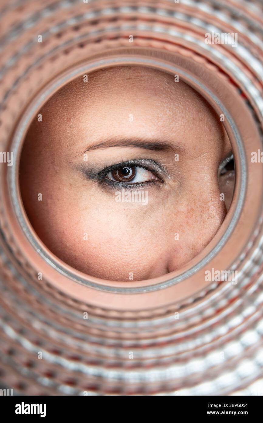 Gros plan de l'œil d'une femme regardant à travers l'ouverture circulaire d'un tuyau texturé, capturant les détails de son maquillage, de la texture de la peau et de la pipe Banque D'Images