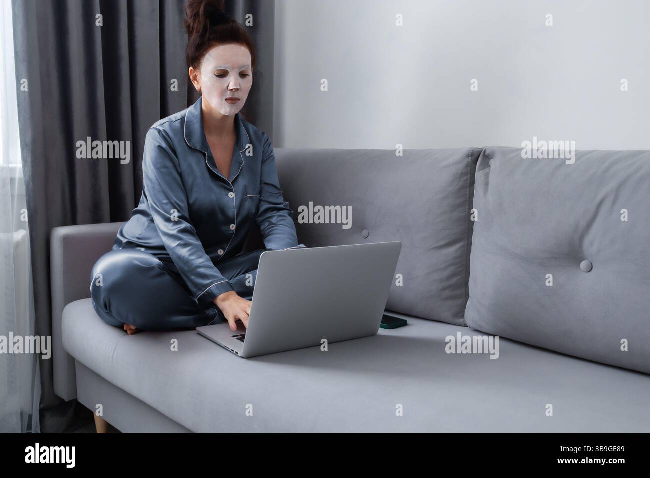 Une femme en pyjama est assise sur un canapé avec un masque facial, travaillant sur un ordinateur portable, incarnant une installation de bureau à domicile confortable et axée sur les soins personnels. Banque D'Images