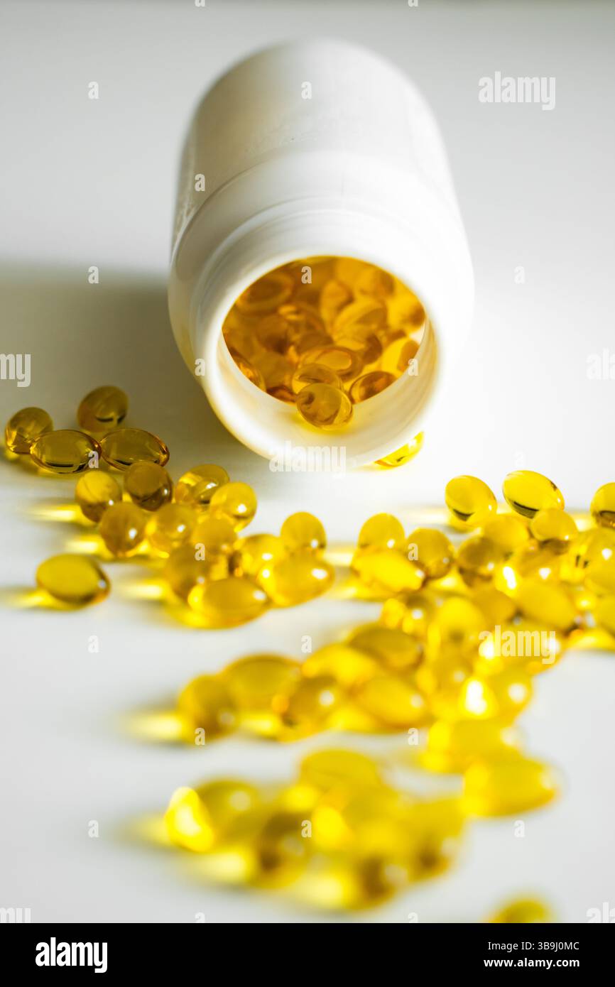 Capsules d'huile de poisson dorée dispersées largement autour d'une bouteille en plastique, illustrant l'apport quotidien de supplément. La lumière vive améliore la texture douce et c Banque D'Images