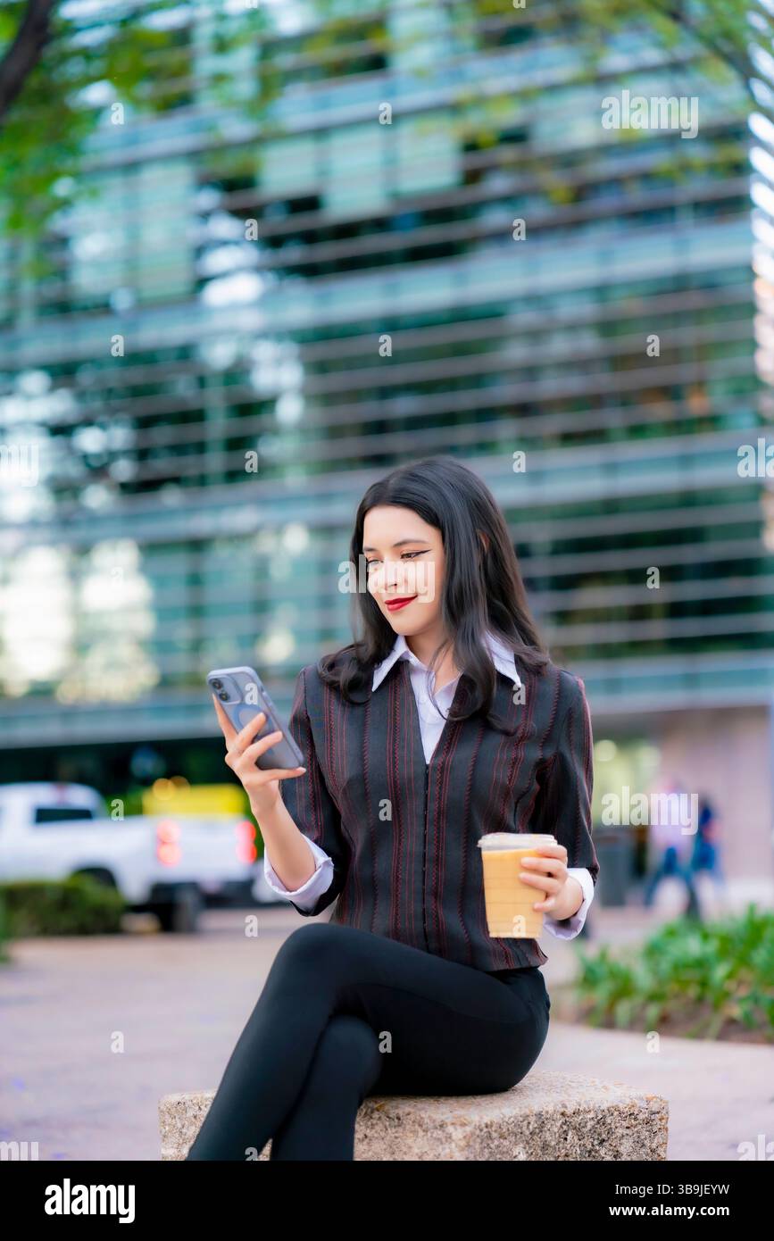 Femme Latina dans une tenue décontractée formelle est assis à l'extérieur sur Reforma, souriant tout en vérifiant son téléphone et tenant un café glacé à Mexico. Banque D'Images