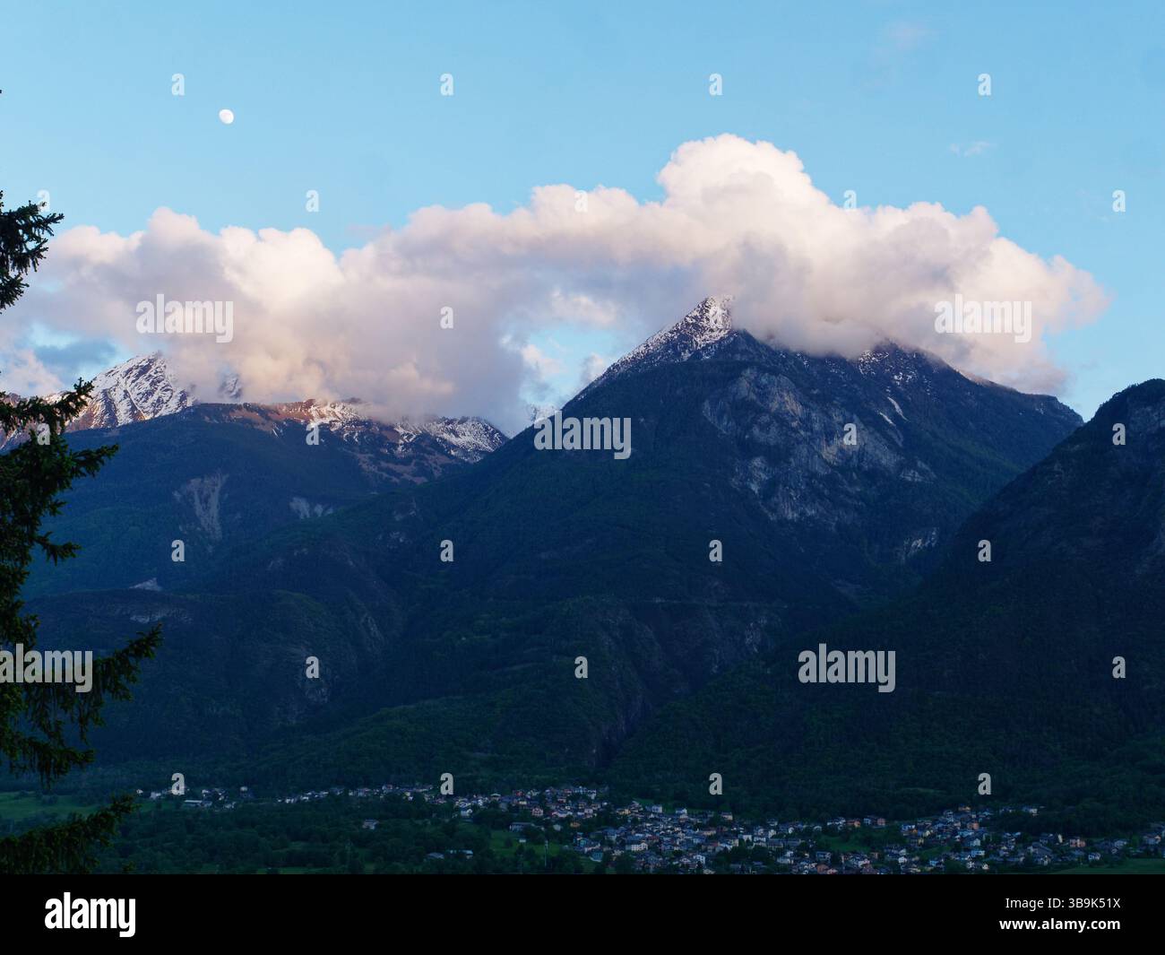 Lune haute dans un ciel bleu au-dessus des nuages blancs, avec des montagnes enneigées et la ville de Fenis ci-dessous. Vallée d'Aoste, NW Italie, 9 mai 2025 Banque D'Images