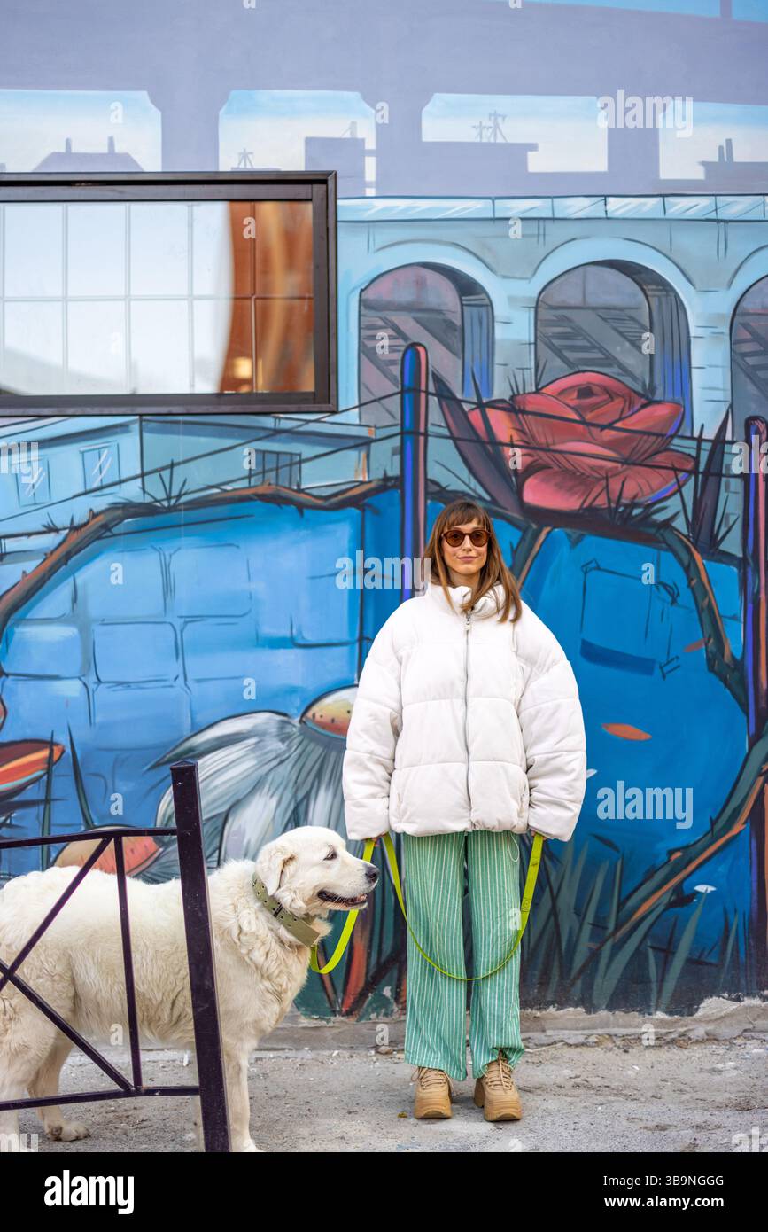 Une femme promène son chien blanc devant une murale de rue vivante dans un quartier romain créatif, mêlant art urbain et moments quotidiens Banque D'Images