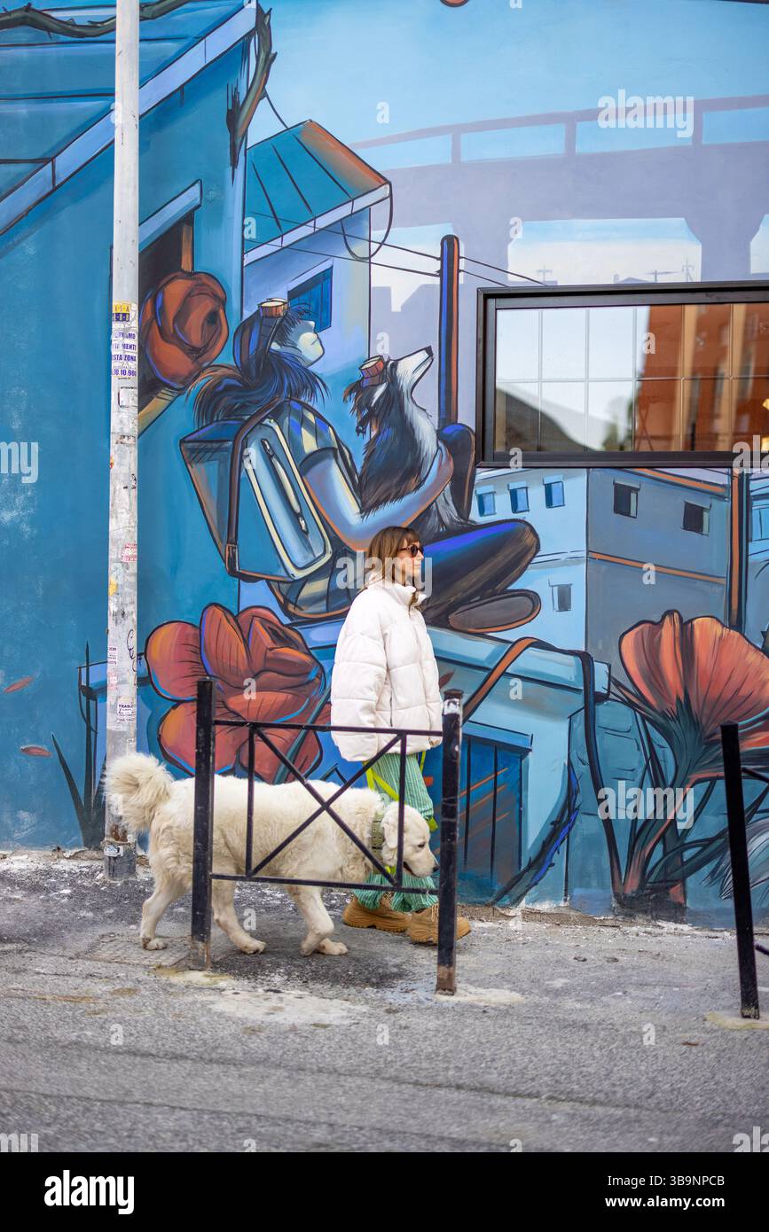 Une femme promène son chien blanc devant une murale de rue vivante dans un quartier romain créatif, mêlant art urbain et moments quotidiens Banque D'Images