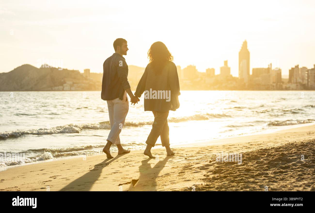 Couple silhouetté profitant d'une promenade romantique sur la plage pendant le coucher du soleil, pieds nus sur le sable avec un panorama de la ville et des montagnes en arrière-plan. La vie Banque D'Images
