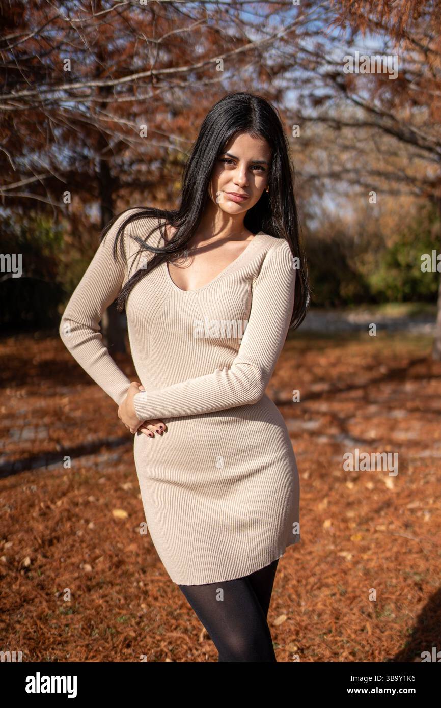 Femme brune aux cheveux longs posant dans un parc en automne, portant une mini robe beige et des collants noirs Banque D'Images