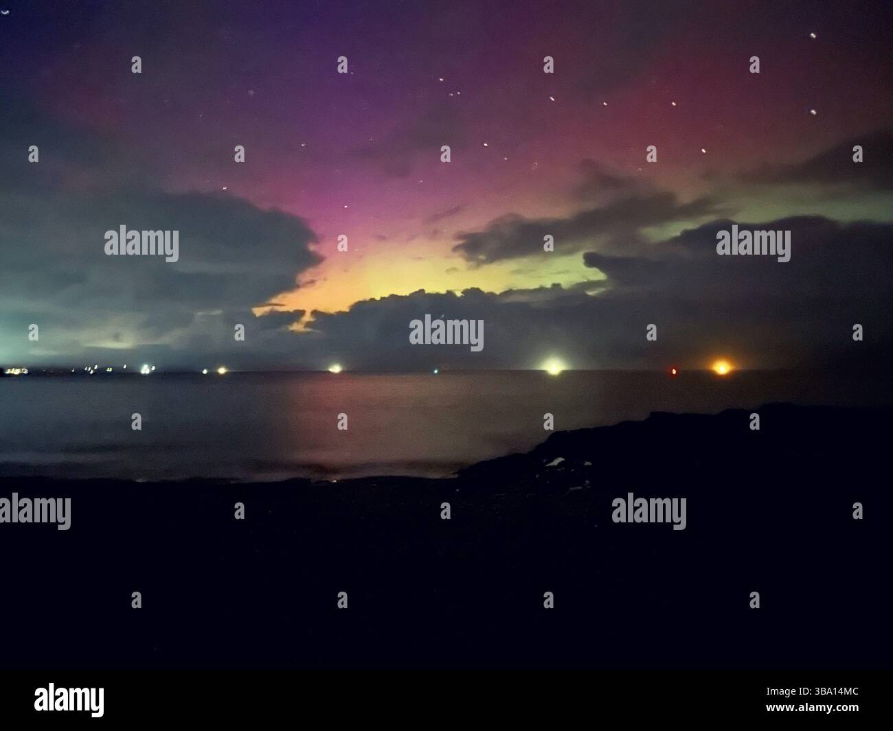 Aurora borealis de Penmon point, Anglesey. Lumières roses, violettes, orange et vertes au-dessus de la plage et de la mer avec lumières artificielles offshore. - Image de stock capturée avec un smartphone