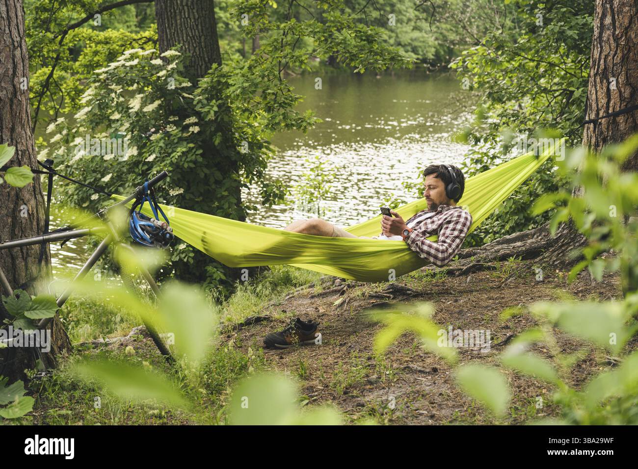 L'homme voyage à vélo, se détendre dans un hamac vert, surfer sur Internet sur un smartphone, écouter de la musique sur des écouteurs dans la forêt près du lac. Cycliste en hammo Banque D'Images