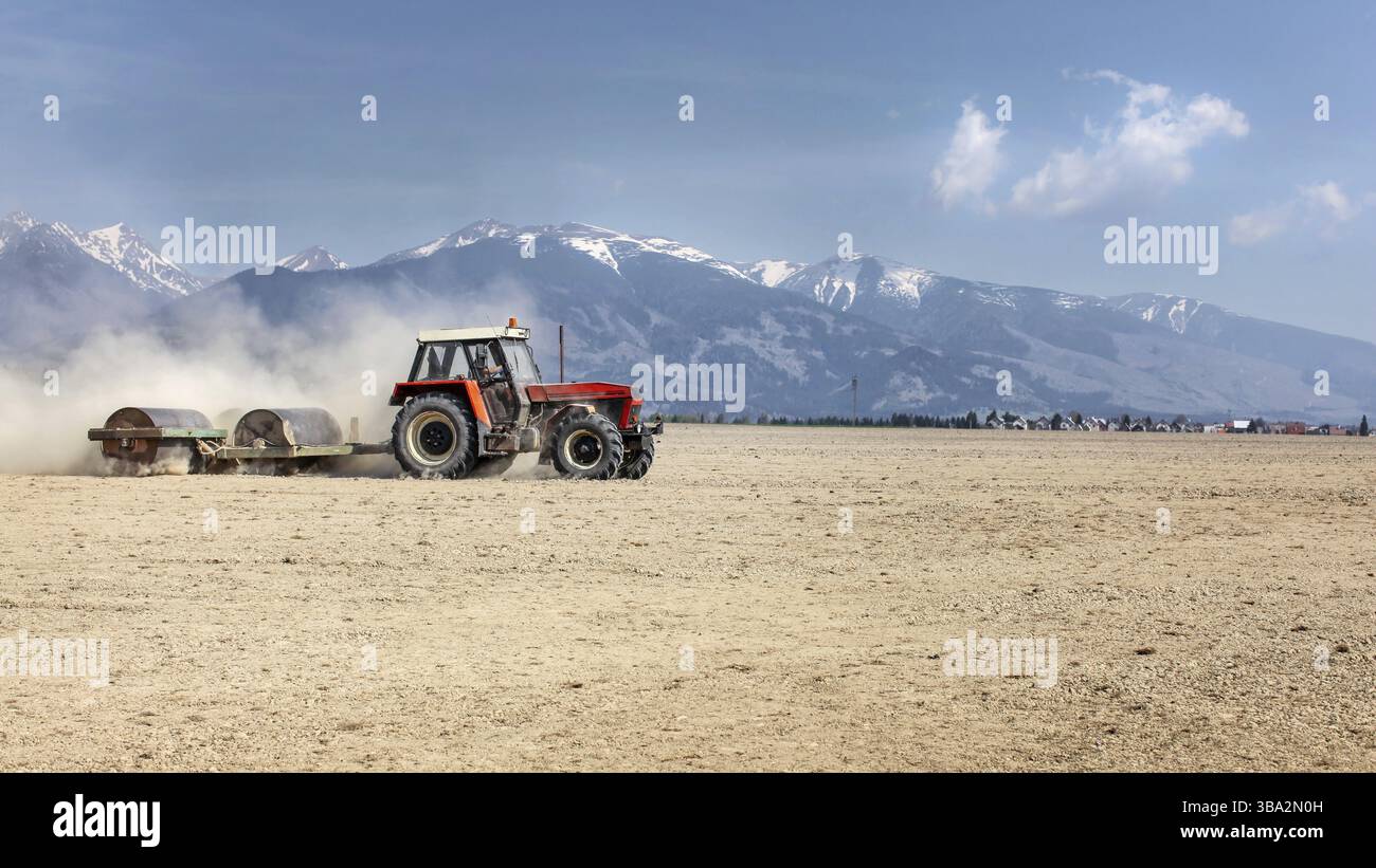 Tracteur tirant un rouleau métallique lourd sur un champ sec sur une belle journée de printemps avec des montagnes en arrière-plan. Préparation du sol, Liptovsky Hradok, Slovaquie, EUR Banque D'Images