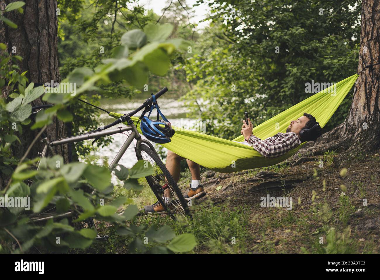 L'homme voyage à vélo, se détendre dans un hamac vert, surfer sur Internet sur un smartphone, écouter de la musique sur des écouteurs dans la forêt près du lac. Cycliste en hammo Banque D'Images