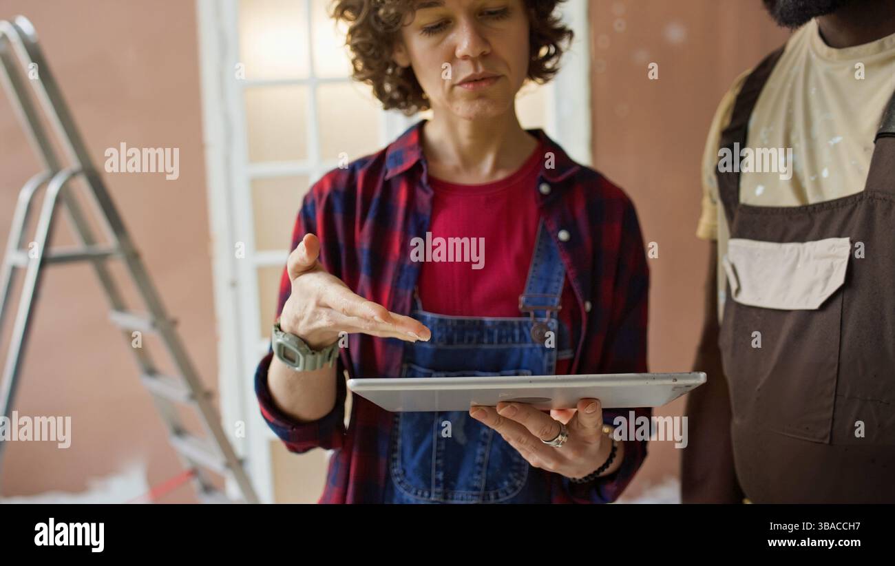 Femme tenant une tablette tout en se tenant debout dans une chambre en rénovation, discutant de plans avec une autre personne, portant des vêtements décontractés, et semblant engagée et concentrée Banque D'Images