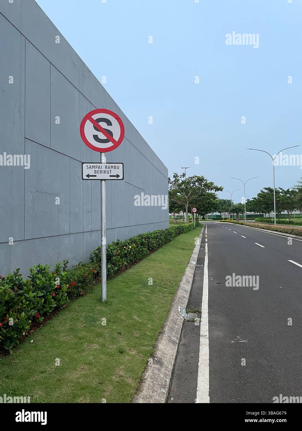 Un panneau de signalisation « No parking » en langue indonésienne, situé dans une rue urbaine propre et moderne avec des plantes soigneusement entretenues et un mur de bâtiment gris. - Image de stock capturée avec un smartphone