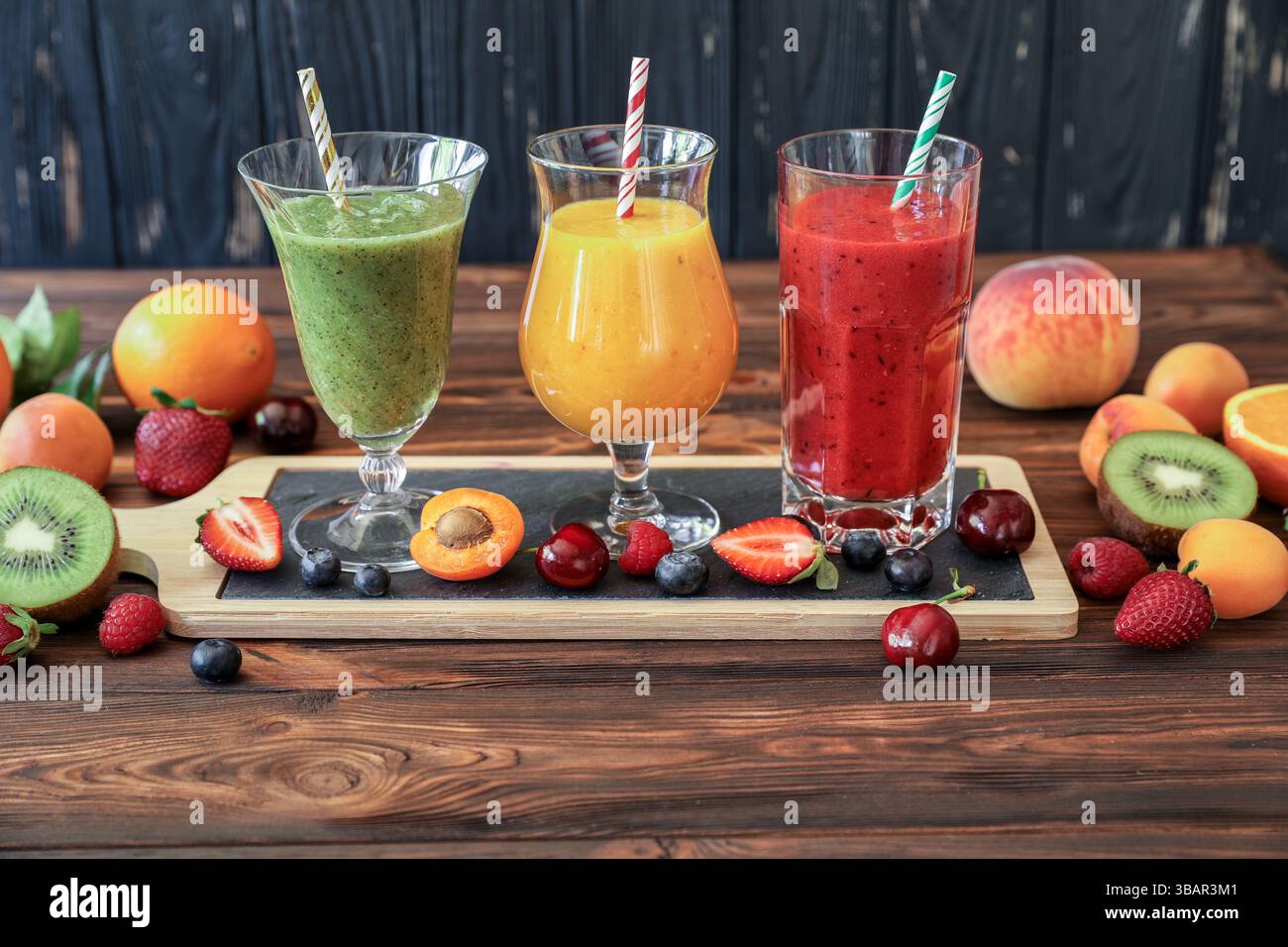 Trois grands verres de smoothies dans les tons vert, jaune et rouge sont assis sur une planche de service en bois. Les fruits environnants comprennent des fraises, des kiwis, des pêches et des oranges, créant une atmosphère rafraîchissante. Banque D'Images