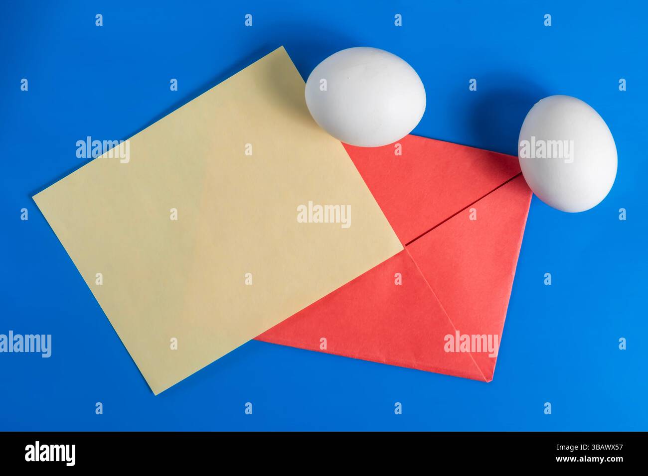 Un arrangement vibrant avec des œufs blancs, une feuille de papier jaune et une enveloppe rouge sur une surface bleu vif. Parfait pour les vacances, Pâques ou la communication Banque D'Images
